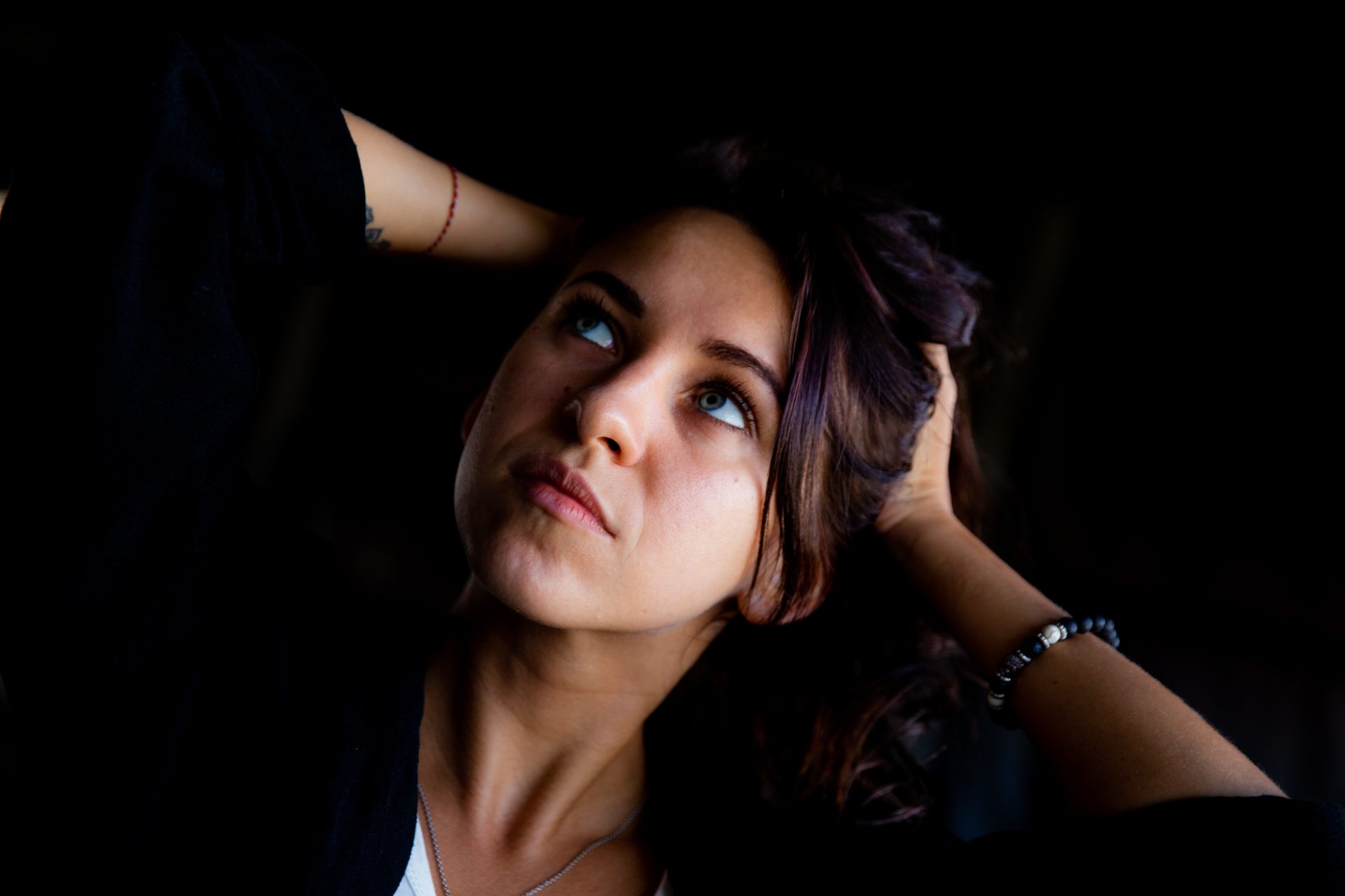 a young woman with purplish brown hair has her hands on her head and is looking up at the sky.