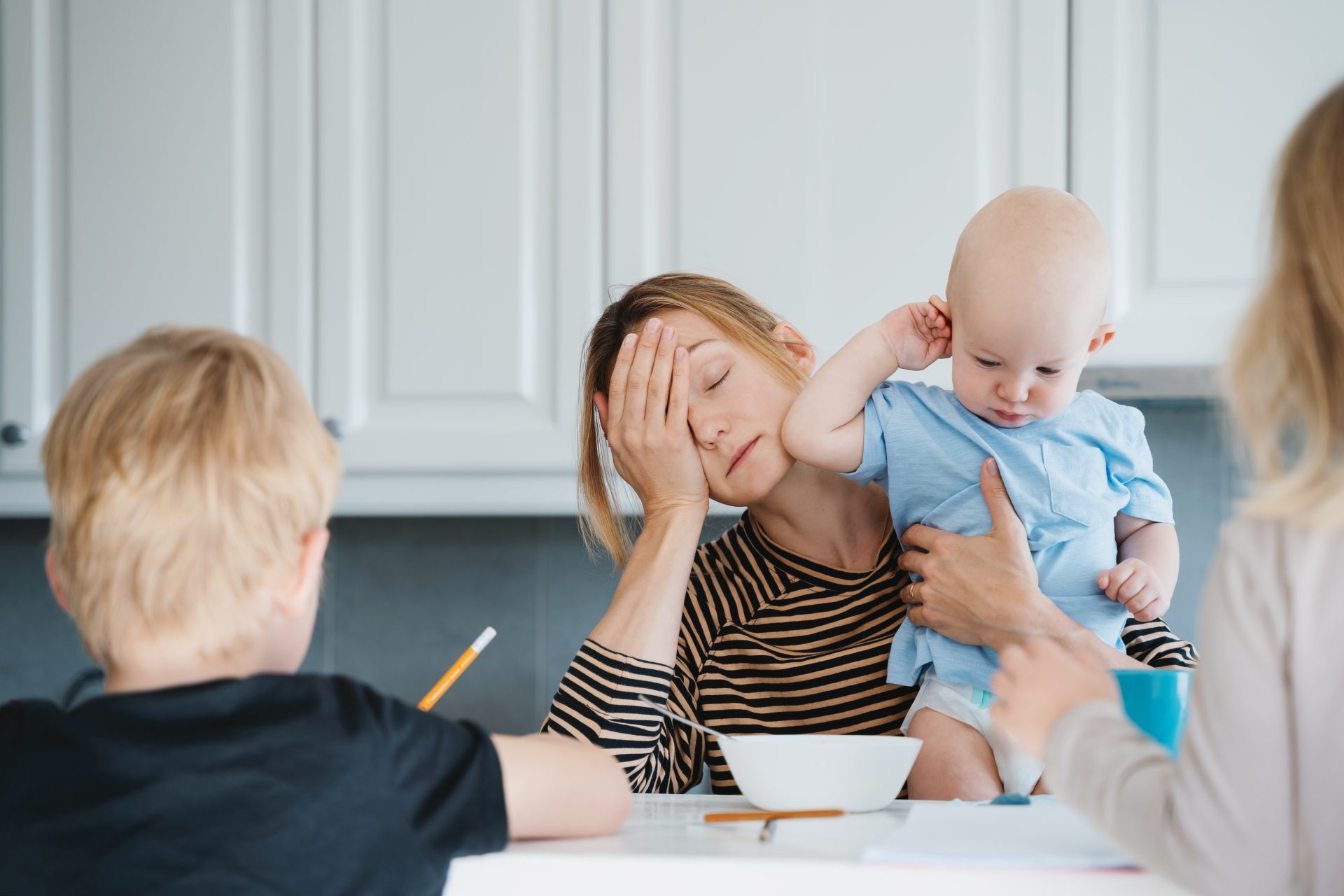 young-exhausted-woman-with-three-children-at-home-2025-03-10-01-20-55-utc a mom with light brown hair is tired, with her head on her hands, holding a bald baby while another young boy (unseen) uses a pencil to complete what appears to be homework of some kind. There is also a bowl in front of the mom as though she is tired from taking care of her kids.